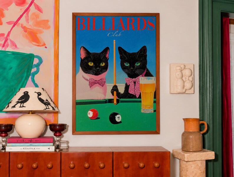 Funny cats playing pool poster featuring two black-faced cats wearing pink patterned bows behind a bright green billiards table. One tuxedo cat has a white body and yellow eyes, while the other is black with green eyes and holds an orange pool cue. Red and black billiard balls marked 8 and 7 sit on the felt beside a glass of golden beer. Large red BILLIARDS lettering, delicate Club script stand out against a vivid blue background. This quirky cat artwork suits game room, home bar, gallery wall.