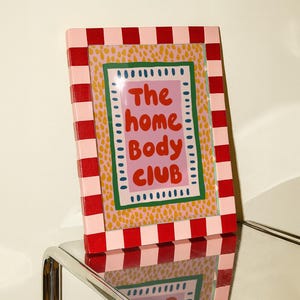 Playful typography poster featuring the phrase The Home Body Club in bold red lettering on a soft pink background. Framed with green and cream borders and surrounded by mustard yellow abstract dots, this colorful maximalist design blends cozy lifestyle humor with eclectic pattern layering and retro-inspired charm.Home Body Poster, Typography Wall Art, Cozy Quote Print, Maximalist Decor, Eclectic Print, Retro Style Poster, Entryway Wall Decor, Bold Letter Art, Trendy Art Print, Dopamine Color Art
