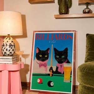 Funny cats playing pool poster featuring two black-faced cats wearing pink patterned bows behind a bright green billiards table. One tuxedo cat has a white body and yellow eyes, while the other is black with green eyes and holds an orange pool cue. Red and black billiard balls marked 8 and 7 sit on the felt beside a glass of golden beer. Large red BILLIARDS lettering, delicate Club script stand out against a vivid blue background. This quirky cat artwork suits game room, home bar, gallery wall.