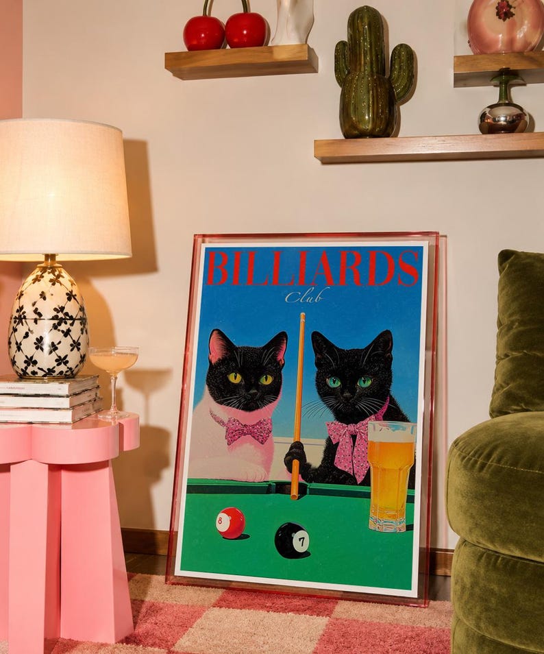 Funny cats playing pool poster featuring two black-faced cats wearing pink patterned bows behind a bright green billiards table. One tuxedo cat has a white body and yellow eyes, while the other is black with green eyes and holds an orange pool cue. Red and black billiard balls marked 8 and 7 sit on the felt beside a glass of golden beer. Large red BILLIARDS lettering, delicate Club script stand out against a vivid blue background. This quirky cat artwork suits game room, home bar, gallery wall.