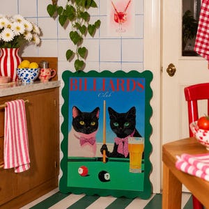 Funny cats playing pool poster featuring two black-faced cats wearing pink patterned bows behind a bright green billiards table. One tuxedo cat has a white body and yellow eyes, while the other is black with green eyes and holds an orange pool cue. Red and black billiard balls marked 8 and 7 sit on the felt beside a glass of golden beer. Large red BILLIARDS lettering, delicate Club script stand out against a vivid blue background. This quirky cat artwork suits game room, home bar, gallery wall.