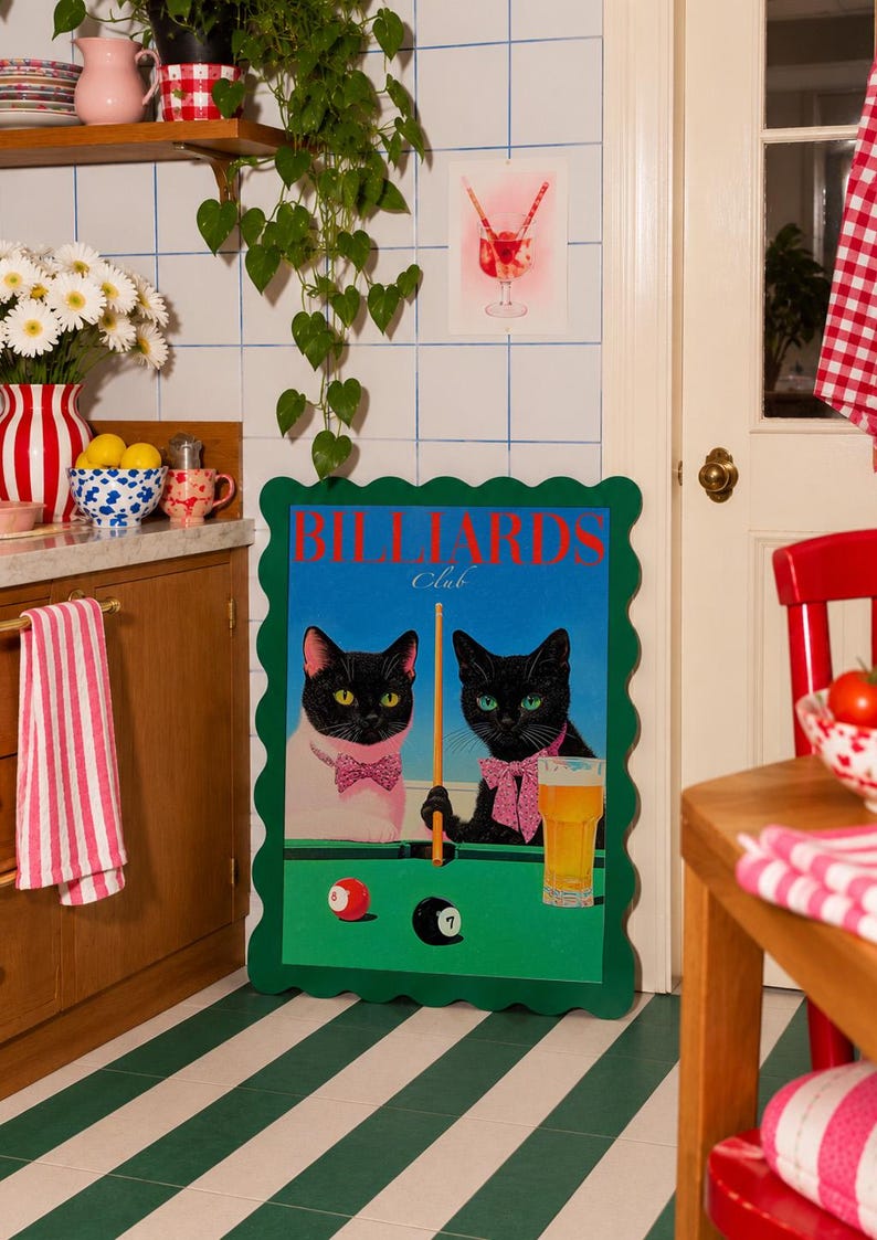 Funny cats playing pool poster featuring two black-faced cats wearing pink patterned bows behind a bright green billiards table. One tuxedo cat has a white body and yellow eyes, while the other is black with green eyes and holds an orange pool cue. Red and black billiard balls marked 8 and 7 sit on the felt beside a glass of golden beer. Large red BILLIARDS lettering, delicate Club script stand out against a vivid blue background. This quirky cat artwork suits game room, home bar, gallery wall.