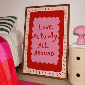 A playful pink and red typography print featuring the hand-lettered message “Love Actually All Around” inside a scalloped frame. The warm peach background, bold lettering and imperfect drawn texture give the artwork a cheerful, handmade character. Designed for bedrooms, living rooms, gallery walls or an above-bed display, this romantic wall art adds color without feeling overly formal. A thoughtful choice for anniversaries, Valentine’s Day, newlyweds or anyone who loves expressive quote posters.