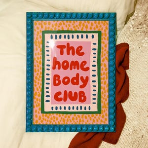 Playful typography poster featuring the phrase The Home Body Club in bold red lettering on a soft pink background. Framed with green and cream borders and surrounded by mustard yellow abstract dots, this colorful maximalist design blends cozy lifestyle humor with eclectic pattern layering and retro-inspired charm.Home Body Poster, Typography Wall Art, Cozy Quote Print, Maximalist Decor, Eclectic Print, Retro Style Poster, Entryway Wall Decor, Bold Letter Art, Trendy Art Print, Dopamine Color Art
