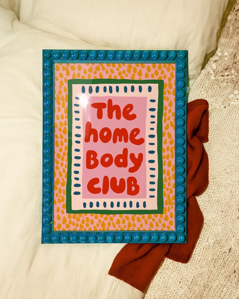 Playful typography poster featuring the phrase The Home Body Club in bold red lettering on a soft pink background. Framed with green and cream borders and surrounded by mustard yellow abstract dots, this colorful maximalist design blends cozy lifestyle humor with eclectic pattern layering and retro-inspired charm.Home Body Poster, Typography Wall Art, Cozy Quote Print, Maximalist Decor, Eclectic Print, Retro Style Poster, Entryway Wall Decor, Bold Letter Art, Trendy Art Print, Dopamine Color Art