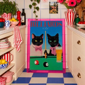 Funny cats playing pool poster featuring two black-faced cats wearing pink patterned bows behind a bright green billiards table. One tuxedo cat has a white body and yellow eyes, while the other is black with green eyes and holds an orange pool cue. Red and black billiard balls marked 8 and 7 sit on the felt beside a glass of golden beer. Large red BILLIARDS lettering, delicate Club script stand out against a vivid blue background. This quirky cat artwork suits game room, home bar, gallery wall.