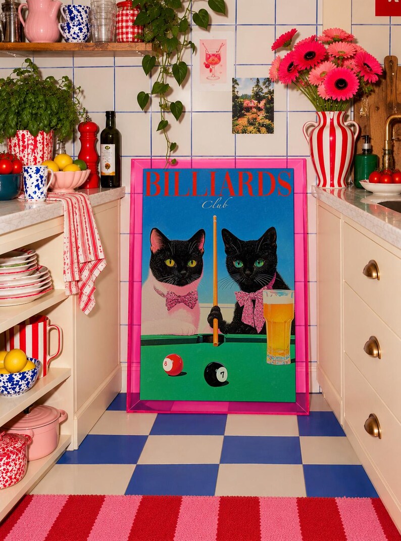 Funny cats playing pool poster featuring two black-faced cats wearing pink patterned bows behind a bright green billiards table. One tuxedo cat has a white body and yellow eyes, while the other is black with green eyes and holds an orange pool cue. Red and black billiard balls marked 8 and 7 sit on the felt beside a glass of golden beer. Large red BILLIARDS lettering, delicate Club script stand out against a vivid blue background. This quirky cat artwork suits game room, home bar, gallery wall.