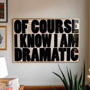 Puede incluir: Una obra de arte enmarcada con el texto "OF COURSE I KNOW I AM DRAMATIC" en letras negras en negrita. El fondo es un collage de páginas de libros antiguos, y el marco es dorado.
