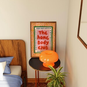 Playful typography poster featuring the phrase The Home Body Club in bold red lettering on a soft pink background. Framed with green and cream borders and surrounded by mustard yellow abstract dots, this colorful maximalist design blends cozy lifestyle humor with eclectic pattern layering and retro-inspired charm.Home Body Poster, Typography Wall Art, Cozy Quote Print, Maximalist Decor, Eclectic Print, Retro Style Poster, Entryway Wall Decor, Bold Letter Art, Trendy Art Print, Dopamine Color Art