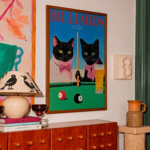 Funny cats playing pool poster featuring two black-faced cats wearing pink patterned bows behind a bright green billiards table. One tuxedo cat has a white body and yellow eyes, while the other is black with green eyes and holds an orange pool cue. Red and black billiard balls marked 8 and 7 sit on the felt beside a glass of golden beer. Large red BILLIARDS lettering, delicate Club script stand out against a vivid blue background. This quirky cat artwork suits game room, home bar, gallery wall.