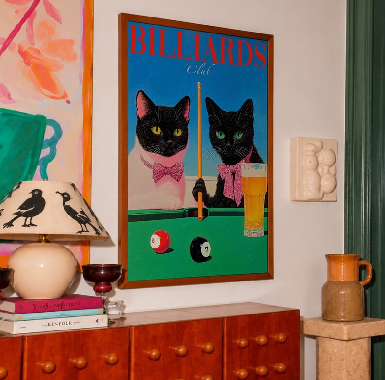 Funny cats playing pool poster featuring two black-faced cats wearing pink patterned bows behind a bright green billiards table. One tuxedo cat has a white body and yellow eyes, while the other is black with green eyes and holds an orange pool cue. Red and black billiard balls marked 8 and 7 sit on the felt beside a glass of golden beer. Large red BILLIARDS lettering, delicate Club script stand out against a vivid blue background. This quirky cat artwork suits game room, home bar, gallery wall.