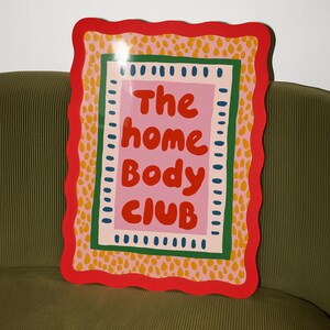 Playful typography poster featuring the phrase The Home Body Club in bold red lettering on a soft pink background. Framed with green and cream borders and surrounded by mustard yellow abstract dots, this colorful maximalist design blends cozy lifestyle humor with eclectic pattern layering and retro-inspired charm.Home Body Poster, Typography Wall Art, Cozy Quote Print, Maximalist Decor, Eclectic Print, Retro Style Poster, Entryway Wall Decor, Bold Letter Art, Trendy Art Print, Dopamine Color Art