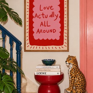 A playful pink and red typography print featuring the hand-lettered message “Love Actually All Around” inside a scalloped frame. The warm peach background, bold lettering and imperfect drawn texture give the artwork a cheerful, handmade character. Designed for bedrooms, living rooms, gallery walls or an above-bed display, this romantic wall art adds color without feeling overly formal. A thoughtful choice for anniversaries, Valentine’s Day, newlyweds or anyone who loves expressive quote posters.