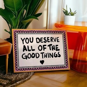 Puede incluir: Un letrero enmarcado con el mensaje "YOU DESERVE ALL OF THE GOOD THINGS" en letras negras, un coraz&oacute;n negro y un borde festoneado rosa y negro. El marco es dorado.