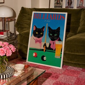 Funny cats playing pool poster featuring two black-faced cats wearing pink patterned bows behind a bright green billiards table. One tuxedo cat has a white body and yellow eyes, while the other is black with green eyes and holds an orange pool cue. Red and black billiard balls marked 8 and 7 sit on the felt beside a glass of golden beer. Large red BILLIARDS lettering, delicate Club script stand out against a vivid blue background. This quirky cat artwork suits game room, home bar, gallery wall.