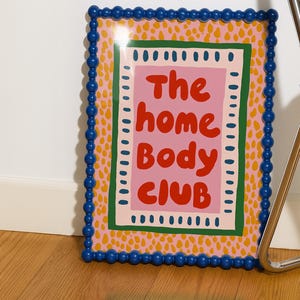 Playful typography poster featuring the phrase The Home Body Club in bold red lettering on a soft pink background. Framed with green and cream borders and surrounded by mustard yellow abstract dots, this colorful maximalist design blends cozy lifestyle humor with eclectic pattern layering and retro-inspired charm.Home Body Poster, Typography Wall Art, Cozy Quote Print, Maximalist Decor, Eclectic Print, Retro Style Poster, Entryway Wall Decor, Bold Letter Art, Trendy Art Print, Dopamine Color Art