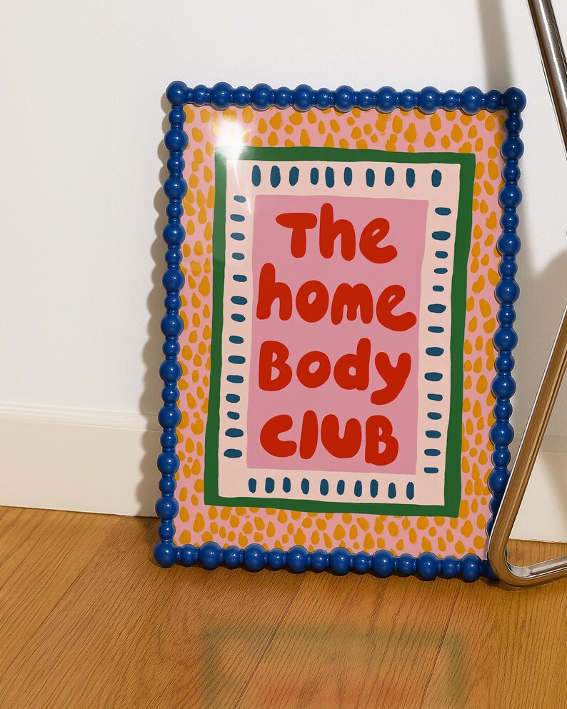 Playful typography poster featuring the phrase The Home Body Club in bold red lettering on a soft pink background. Framed with green and cream borders and surrounded by mustard yellow abstract dots, this colorful maximalist design blends cozy lifestyle humor with eclectic pattern layering and retro-inspired charm.Home Body Poster, Typography Wall Art, Cozy Quote Print, Maximalist Decor, Eclectic Print, Retro Style Poster, Entryway Wall Decor, Bold Letter Art, Trendy Art Print, Dopamine Color Art