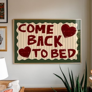 Puede incluir: Arte mural enmarcado con el texto "COME BACK TO BED" en letras rojo oscuro. El fondo tiene rayas verticales beige y dos corazones rojo oscuro. El marco es dorado con un borde verde.