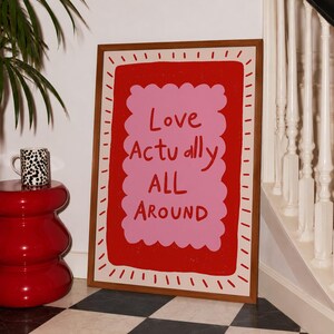 A playful pink and red typography print featuring the hand-lettered message “Love Actually All Around” inside a scalloped frame. The warm peach background, bold lettering and imperfect drawn texture give the artwork a cheerful, handmade character. Designed for bedrooms, living rooms, gallery walls or an above-bed display, this romantic wall art adds color without feeling overly formal. A thoughtful choice for anniversaries, Valentine’s Day, newlyweds or anyone who loves expressive quote posters.