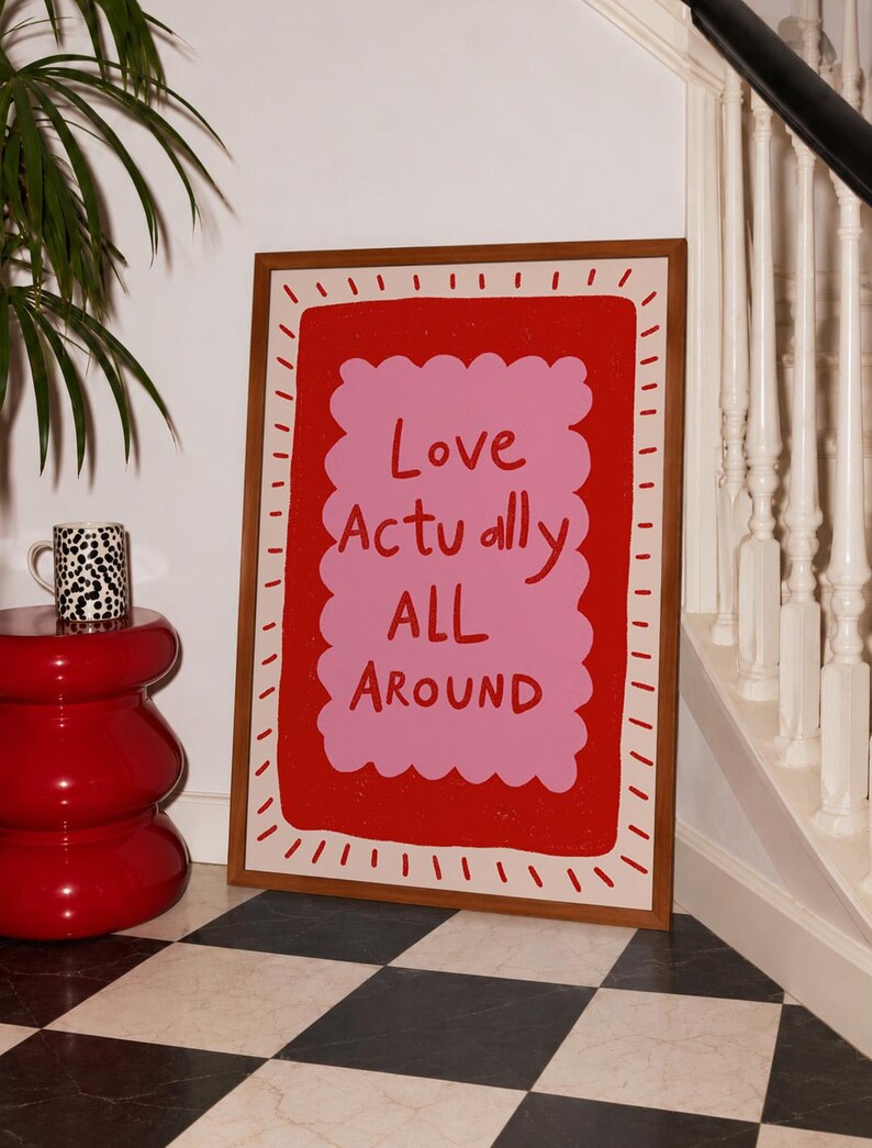 A playful pink and red typography print featuring the hand-lettered message “Love Actually All Around” inside a scalloped frame. The warm peach background, bold lettering and imperfect drawn texture give the artwork a cheerful, handmade character. Designed for bedrooms, living rooms, gallery walls or an above-bed display, this romantic wall art adds color without feeling overly formal. A thoughtful choice for anniversaries, Valentine’s Day, newlyweds or anyone who loves expressive quote posters.