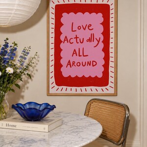A playful pink and red typography print featuring the hand-lettered message “Love Actually All Around” inside a scalloped frame. The warm peach background, bold lettering and imperfect drawn texture give the artwork a cheerful, handmade character. Designed for bedrooms, living rooms, gallery walls or an above-bed display, this romantic wall art adds color without feeling overly formal. A thoughtful choice for anniversaries, Valentine’s Day, newlyweds or anyone who loves expressive quote posters.