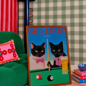 Funny cats playing pool poster featuring two black-faced cats wearing pink patterned bows behind a bright green billiards table. One tuxedo cat has a white body and yellow eyes, while the other is black with green eyes and holds an orange pool cue. Red and black billiard balls marked 8 and 7 sit on the felt beside a glass of golden beer. Large red BILLIARDS lettering, delicate Club script stand out against a vivid blue background. This quirky cat artwork suits game room, home bar, gallery wall.