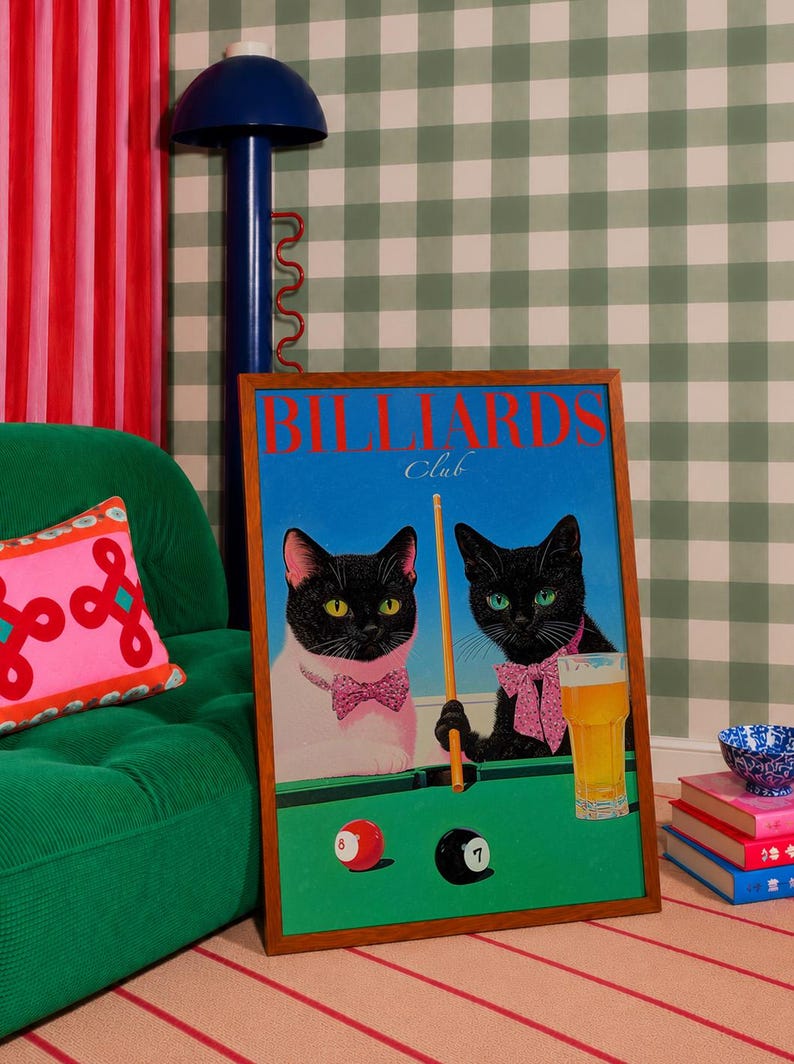 Funny cats playing pool poster featuring two black-faced cats wearing pink patterned bows behind a bright green billiards table. One tuxedo cat has a white body and yellow eyes, while the other is black with green eyes and holds an orange pool cue. Red and black billiard balls marked 8 and 7 sit on the felt beside a glass of golden beer. Large red BILLIARDS lettering, delicate Club script stand out against a vivid blue background. This quirky cat artwork suits game room, home bar, gallery wall.