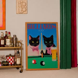 Funny cats playing pool poster featuring two black-faced cats wearing pink patterned bows behind a bright green billiards table. One tuxedo cat has a white body and yellow eyes, while the other is black with green eyes and holds an orange pool cue. Red and black billiard balls marked 8 and 7 sit on the felt beside a glass of golden beer. Large red BILLIARDS lettering, delicate Club script stand out against a vivid blue background. This quirky cat artwork suits game room, home bar, gallery wall.
