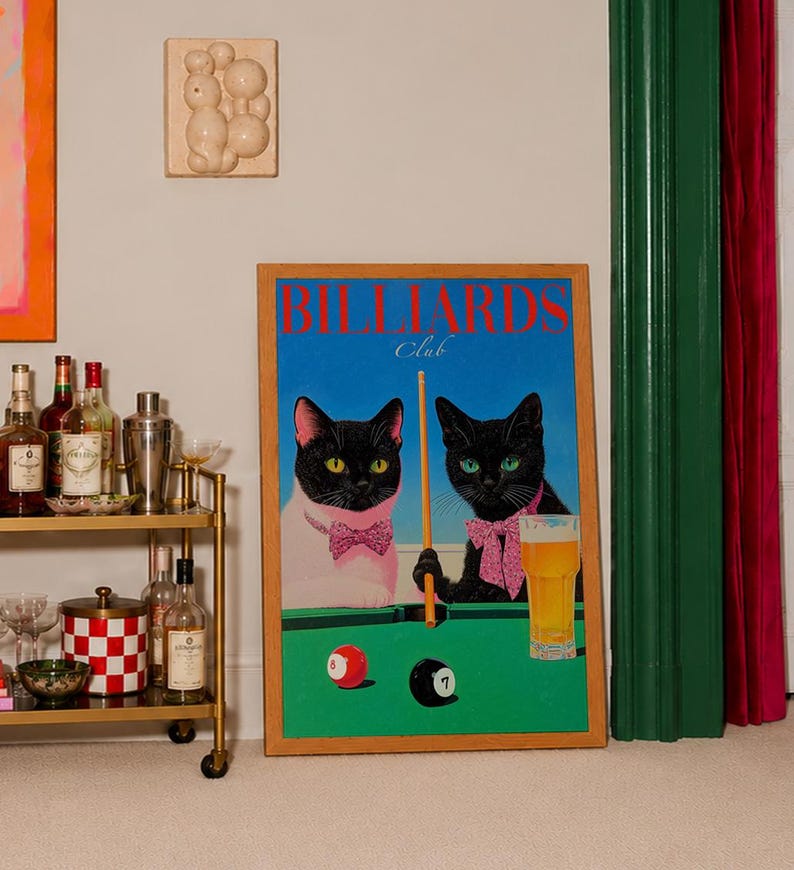 Funny cats playing pool poster featuring two black-faced cats wearing pink patterned bows behind a bright green billiards table. One tuxedo cat has a white body and yellow eyes, while the other is black with green eyes and holds an orange pool cue. Red and black billiard balls marked 8 and 7 sit on the felt beside a glass of golden beer. Large red BILLIARDS lettering, delicate Club script stand out against a vivid blue background. This quirky cat artwork suits game room, home bar, gallery wall.