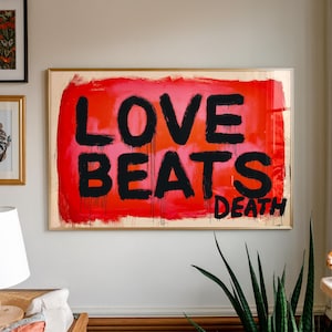 May include: A framed artwork with the words "LOVE BEATS DEATH" in bold black letters against a vibrant red and pink background. The artwork has a gold frame and is hanging on a white wall.