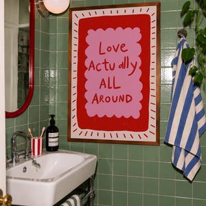 A playful pink and red typography print featuring the hand-lettered message “Love Actually All Around” inside a scalloped frame. The warm peach background, bold lettering and imperfect drawn texture give the artwork a cheerful, handmade character. Designed for bedrooms, living rooms, gallery walls or an above-bed display, this romantic wall art adds color without feeling overly formal. A thoughtful choice for anniversaries, Valentine’s Day, newlyweds or anyone who loves expressive quote posters.