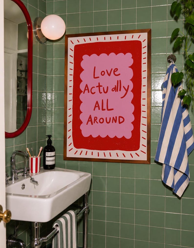 A playful pink and red typography print featuring the hand-lettered message “Love Actually All Around” inside a scalloped frame. The warm peach background, bold lettering and imperfect drawn texture give the artwork a cheerful, handmade character. Designed for bedrooms, living rooms, gallery walls or an above-bed display, this romantic wall art adds color without feeling overly formal. A thoughtful choice for anniversaries, Valentine’s Day, newlyweds or anyone who loves expressive quote posters.