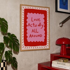 A playful pink and red typography print featuring the hand-lettered message “Love Actually All Around” inside a scalloped frame. The warm peach background, bold lettering and imperfect drawn texture give the artwork a cheerful, handmade character. Designed for bedrooms, living rooms, gallery walls or an above-bed display, this romantic wall art adds color without feeling overly formal. A thoughtful choice for anniversaries, Valentine’s Day, newlyweds or anyone who loves expressive quote posters.