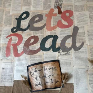 Puede incluir: Una decoración de pared con el texto "let's READ" en grandes letras cursivas de color verde oscuro y terracota sobre un fondo de páginas de libro superpuestas. Debajo, un cartel rectangular muestra el texto "Between the pages of a book is a lovely place to be" con tallos de hierba seca a cada lado.