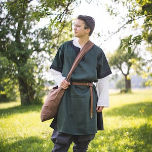 May include: A person wearing a green tunic with a brown leather belt and a brown leather bag slung over their shoulder. They are standing in a grassy field with trees in the background.