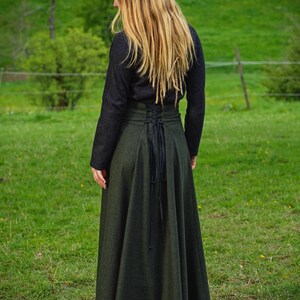 May include: A dark green, long skirt with a high-waisted, corset-style bodice and a flowing A-line shape. The skirt is paired with a black long-sleeved top. The back of the skirt features a black lace-up detail. The person is standing in a grassy field.