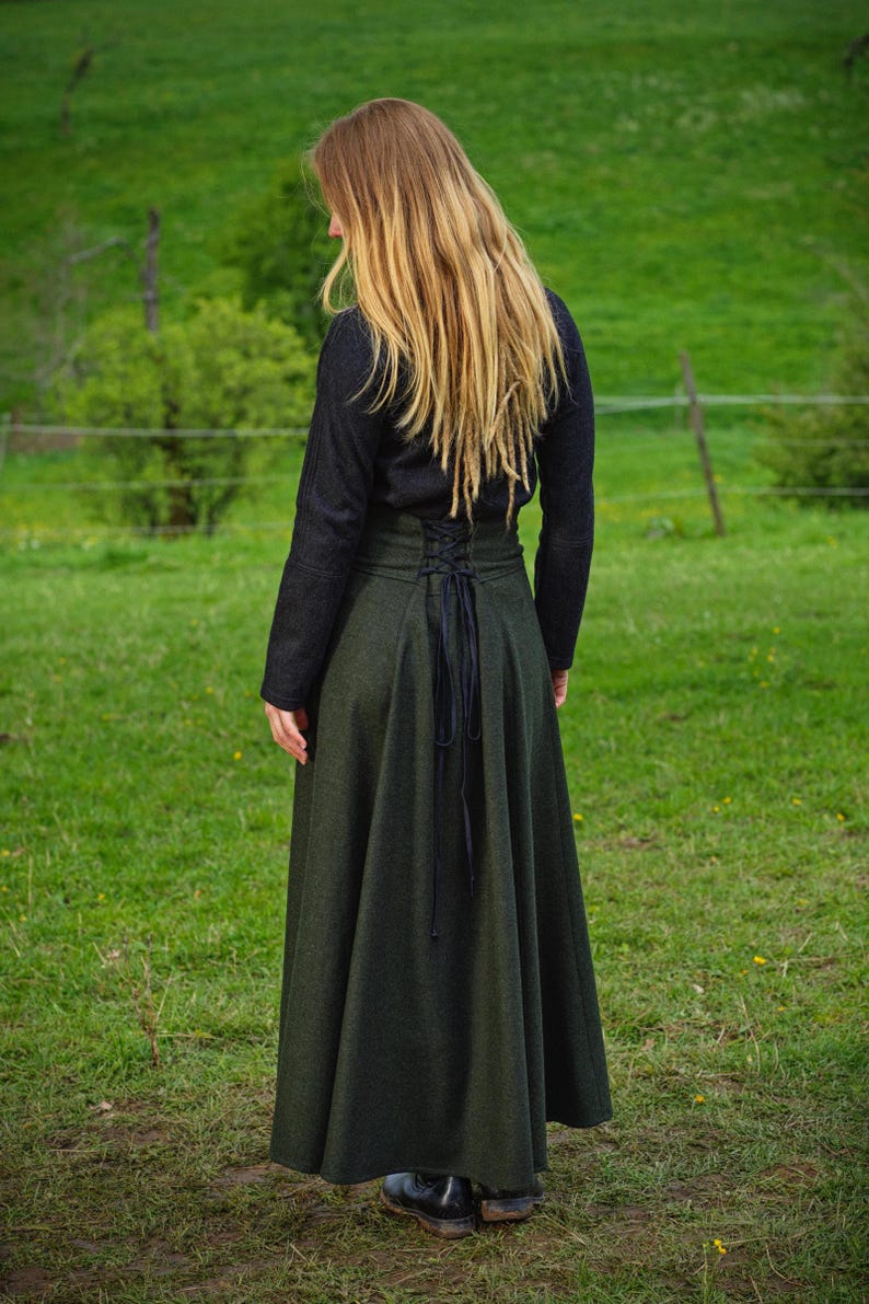 May include: A dark green, long skirt with a high-waisted, corset-style bodice and a flowing A-line shape. The skirt is paired with a black long-sleeved top. The back of the skirt features a black lace-up detail. The person is standing in a grassy field.