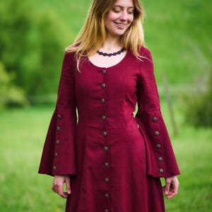 以下が含まれることがあります： ボタン留めのフロントとベルスリーブが付いた、長いワインレッドのドレスを着た女性。ドレスはシンプルでエレガントなデザインで、柔らかく流れるような生地で作られています。