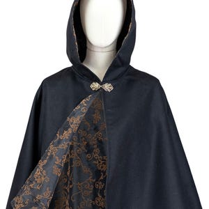 May include: A black hooded cape featuring a gold-toned metal leaf-shaped clasp at the neckline. The interior lining is black with a repeating copper-colored floral scroll pattern that is visible where the front of the cape is open.