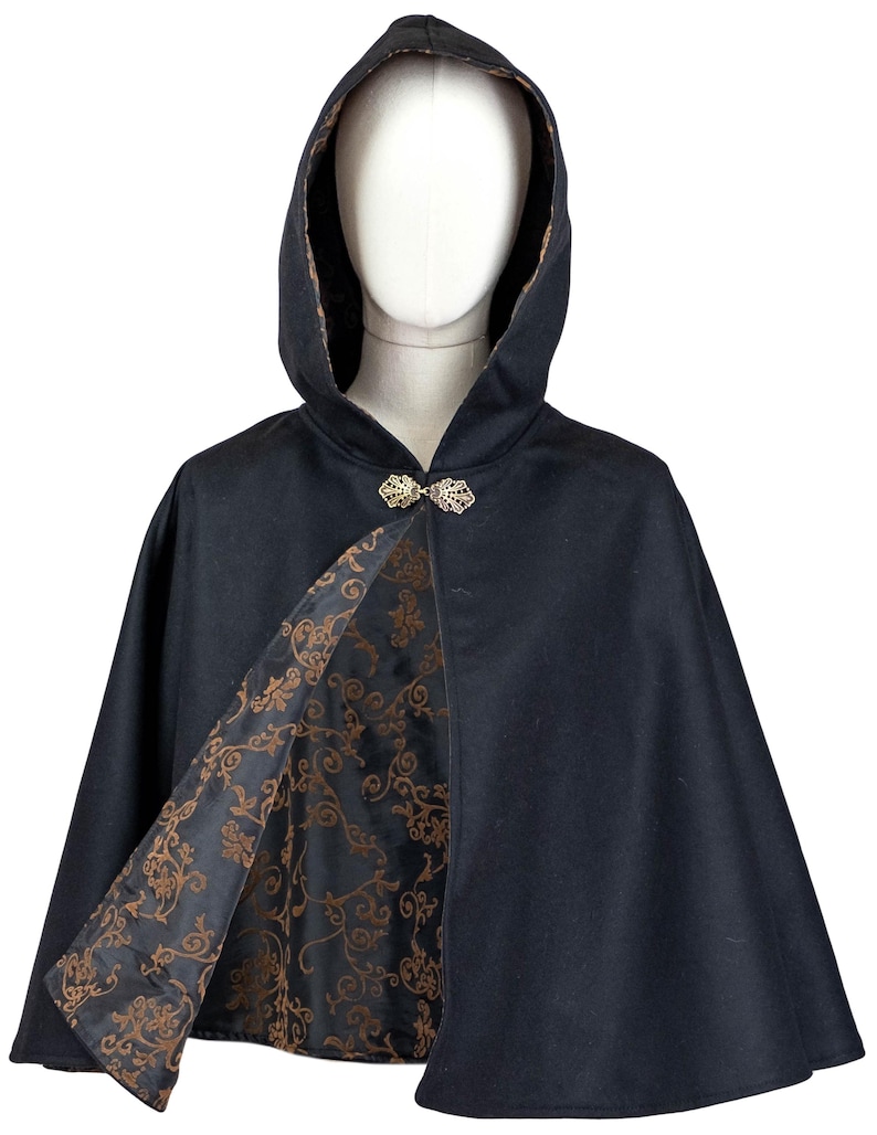 May include: A black hooded cape featuring a gold-toned metal leaf-shaped clasp at the neckline. The interior lining is black with a repeating copper-colored floral scroll pattern that is visible where the front of the cape is open.
