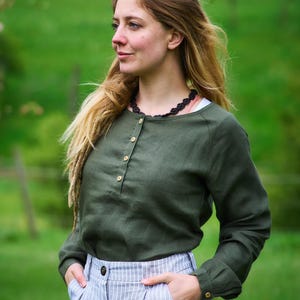 Könnte beinhalten: Ein olivgrünes Langarmhemd mit Henley-Ausschnitt und Holzknöpfen. Das Hemd wird mit hellblau-weiß gestreiften Hosen kombiniert. Eine schwarze Halskette ist sichtbar. Der Hintergrund ist ein grünes Feld.