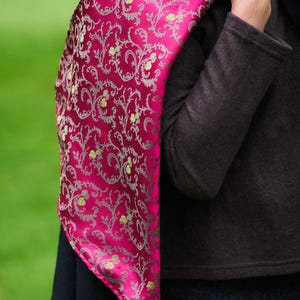 May include: A vibrant magenta scarf featuring a detailed silver filigree pattern with small yellow floral accents. The fabric has a smooth, reflective sheen and is draped over a dark charcoal coat, showcasing its intricate, repeating design.