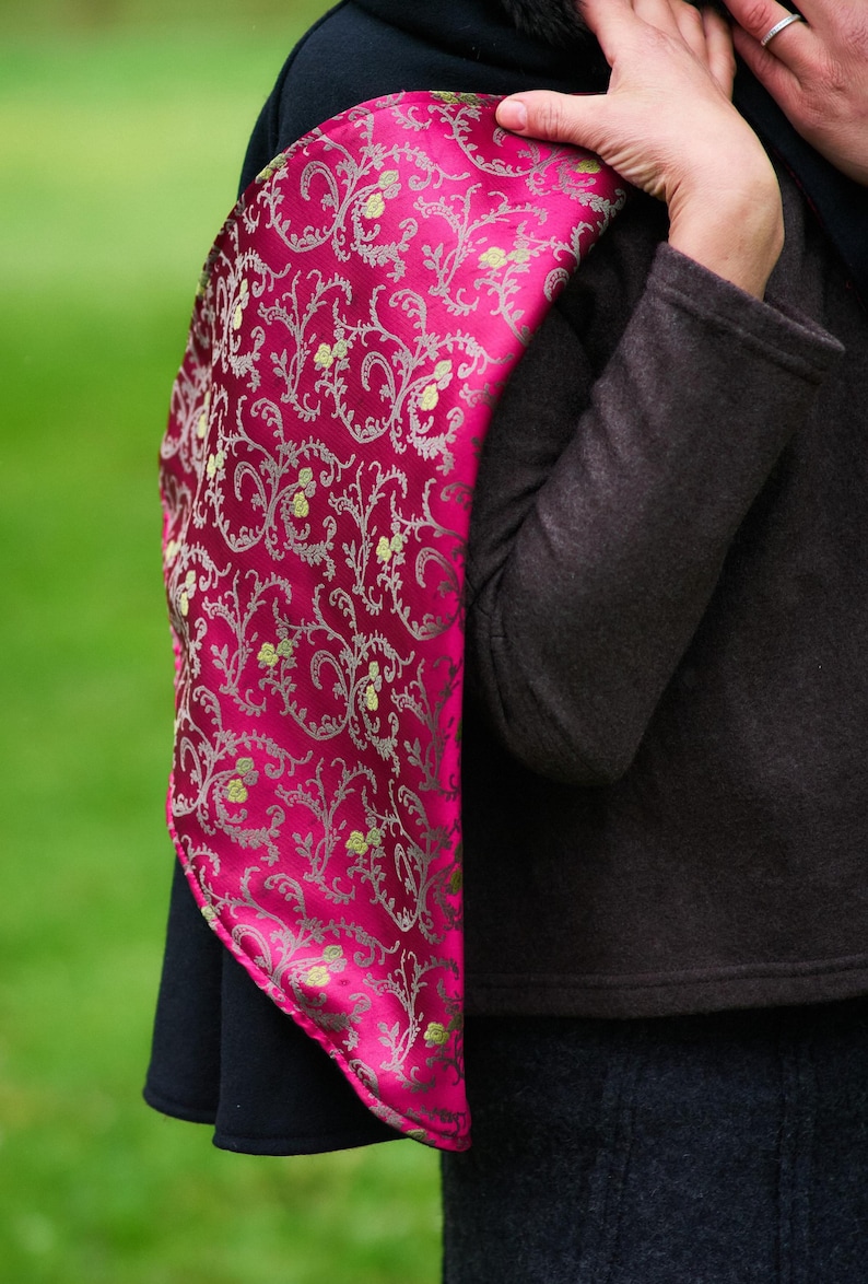 May include: A vibrant magenta scarf featuring a detailed silver filigree pattern with small yellow floral accents. The fabric has a smooth, reflective sheen and is draped over a dark charcoal coat, showcasing its intricate, repeating design.