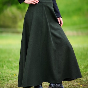 May include: A dark green, full-length skirt with a high waist. The skirt is made of a heavy fabric and flares out from the waist. The person is wearing a black long-sleeved shirt and black boots. The background is a grassy field.