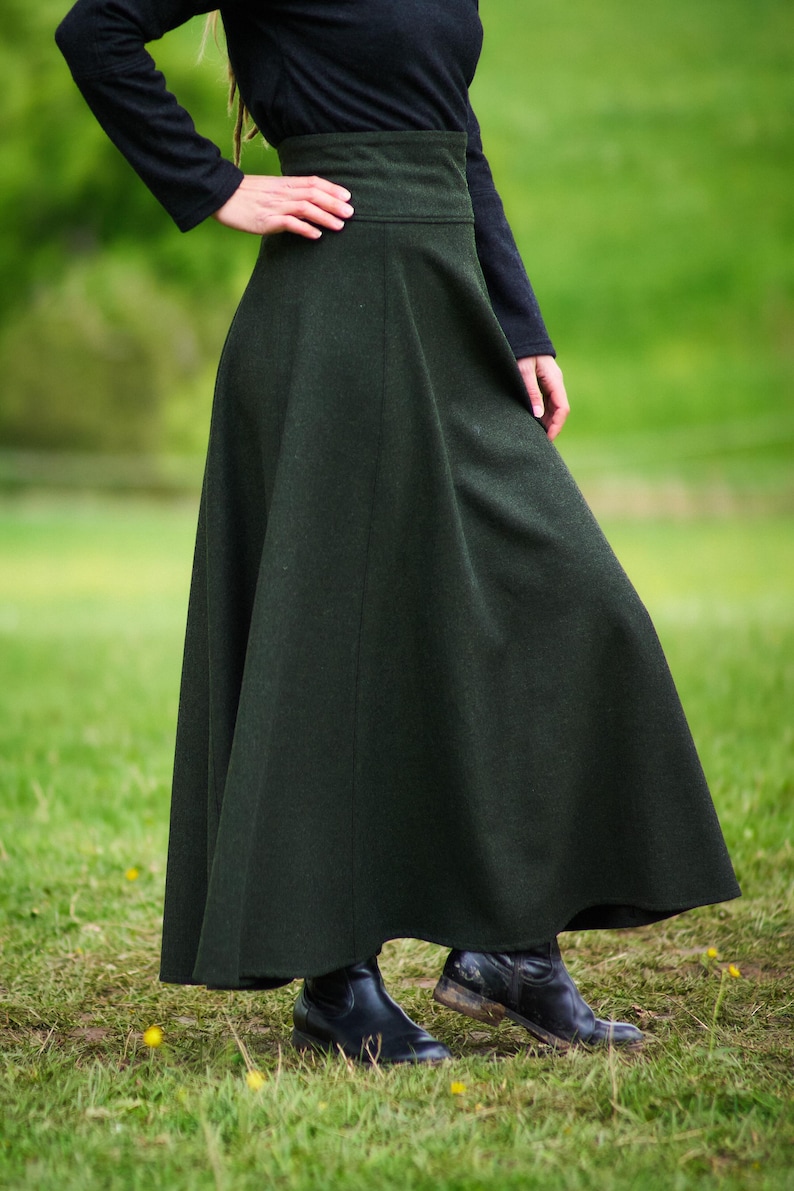 May include: A dark green, full-length skirt with a high waist. The skirt is made of a heavy fabric and flares out from the waist. The person is wearing a black long-sleeved shirt and black boots. The background is a grassy field.