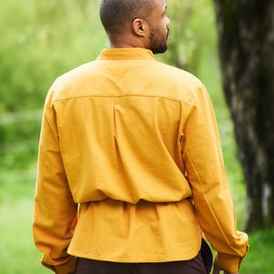 Op de afbeelding: Een persoon draagt een mosterdgeel overhemd met lange mouwen, een bruine riem en een bruine broek. De persoon staat in een grasveld met bomen op de achtergrond.