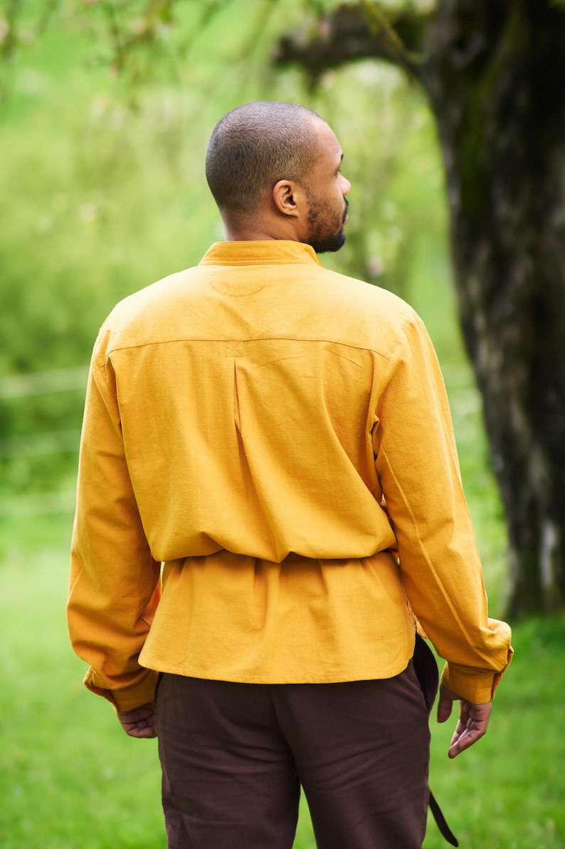 Op de afbeelding: Een persoon draagt een mosterdgeel overhemd met lange mouwen, een bruine riem en een bruine broek. De persoon staat in een grasveld met bomen op de achtergrond.
