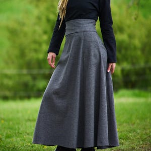 May include: A long, gray skirt with a high waistband paired with a black long-sleeve top. The skirt is floor-length and has a flowing silhouette. The outfit is completed with black boots. The background is a blurred green field.