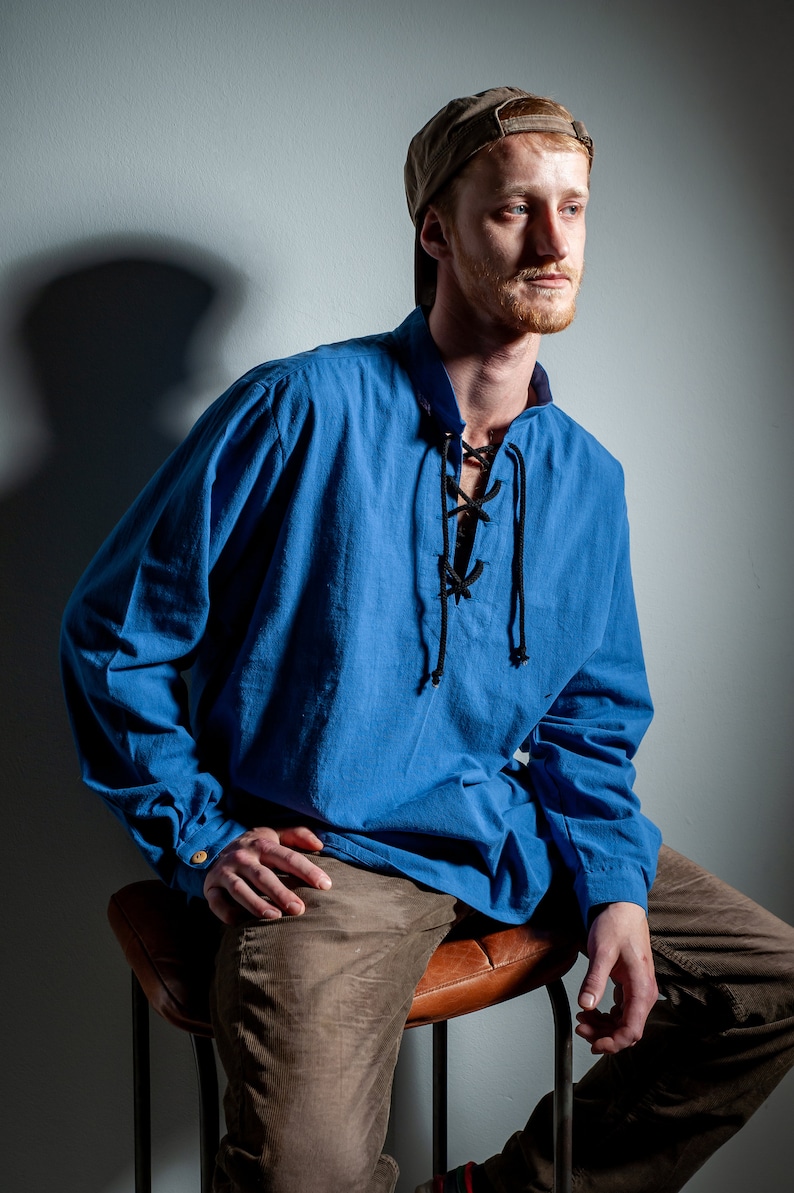Op de afbeelding: Een man draagt een blauw lang mouw shirt met een zwarte veterkraag, een bruine broek en een bruine baseballpet. Hij zit op een bruine leren kruk.