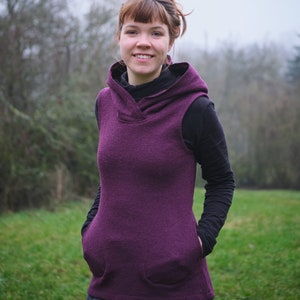 Op de afbeelding: Een vrouw draagt een paarse vest met capuchon met zakken en een zwarte rok. Ze staat in een grasveld met bomen op de achtergrond.